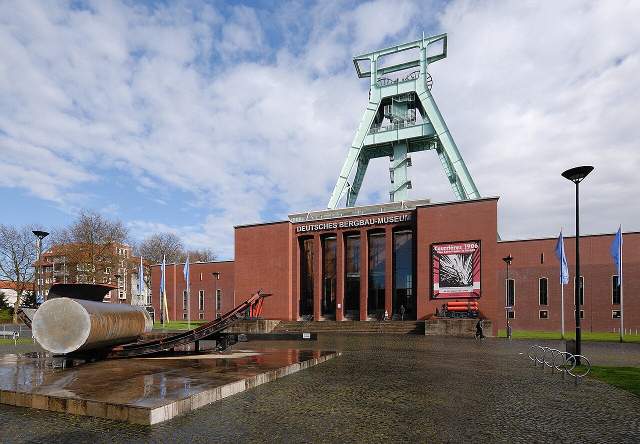 Vor Ihnen steht ein Gebäude aus dunkelrotem Backstein, über dem ein hoher, grüner Förderturm aus Stahlstreben aufragt. In der Mitte liegt ein hohes Glasportal mit schmalen Pfeilern unter einem Schriftband. Die Seitenflügel sind niedriger und haben Fensterreihen. Links im Vorfeld liegen ein großer Stahlzylinder und eine Schiene. Der Vorplatz ist gepflastert.