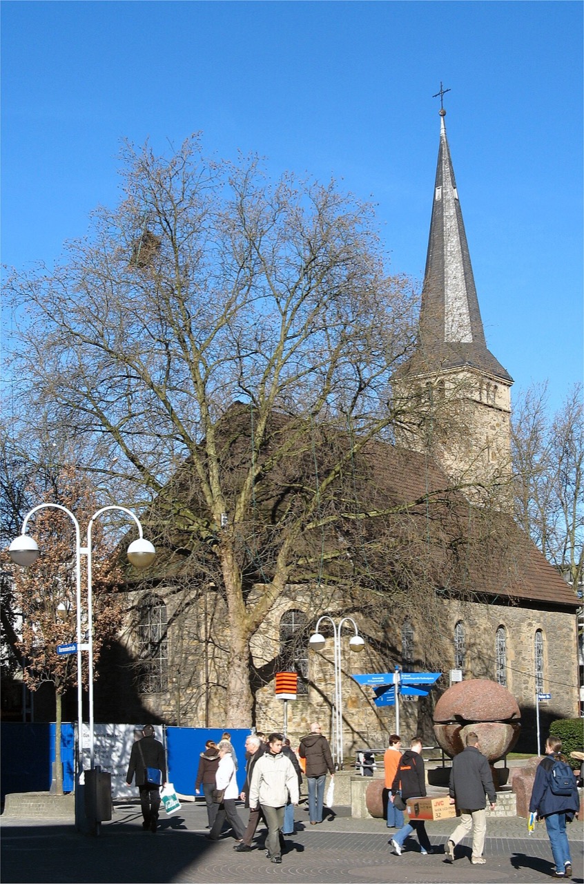 Eine Kirche aus dunklem, verwittertem Bruchstein mit dunklem Dach. Rechts steht ein Turm mit hohem, spitzem Helm aus dunklem Schiefer, der ein Kreuz trägt. Die Fenster des Schiffs sind hoch und schmal. Ein großer Baum verdeckt einen Teil davon. Davor liegt ein gepflasterter Platz mit Laternen, rechts eine runde Steinplastik.