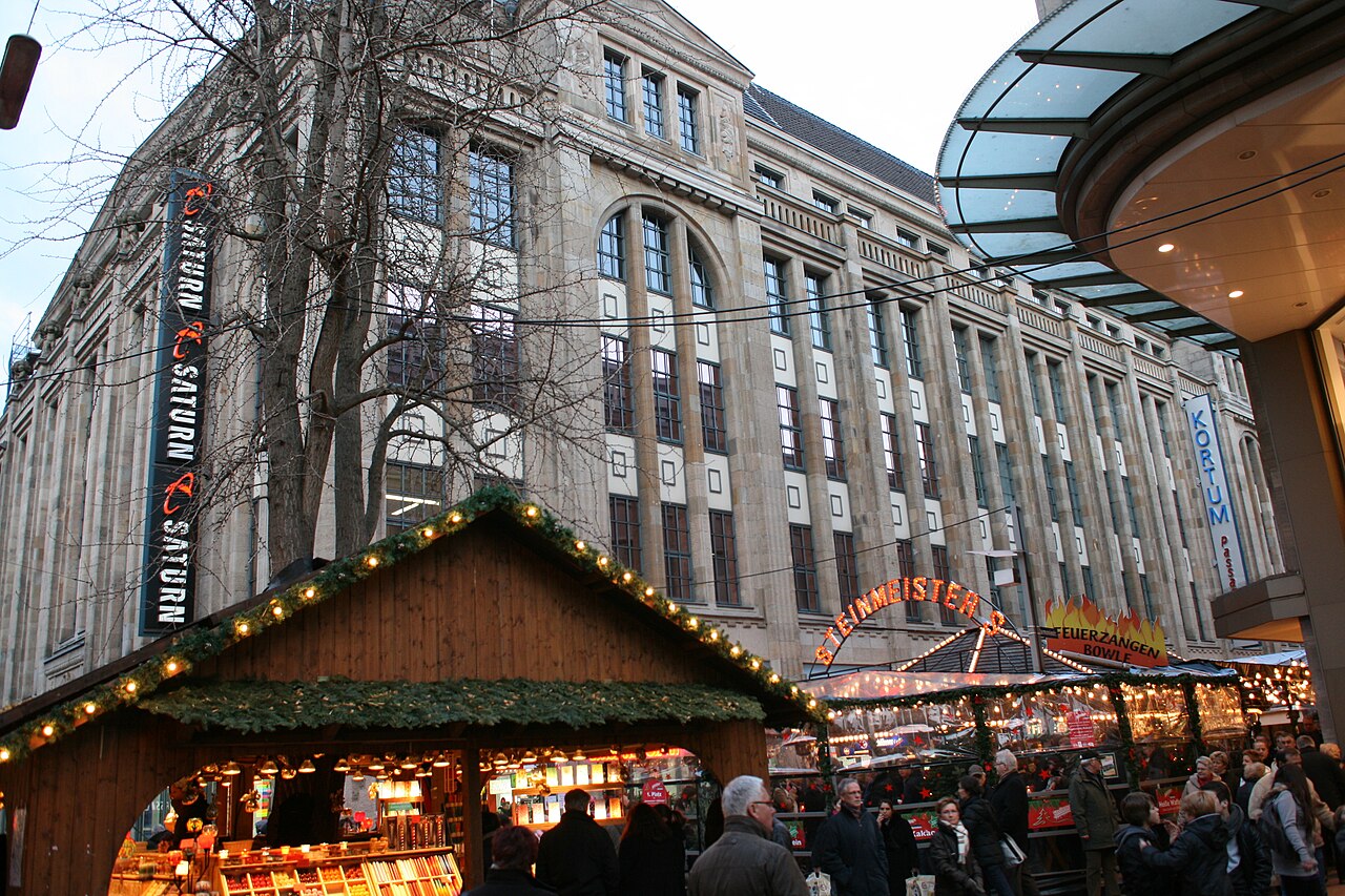 Ein großes Kaufhaus mit heller Natursteinfassade. Senkrechte Steinpfeiler trennen hohe, schmale Fensterfelder, die sich über mehrere Geschosse ziehen. Die Fassade erstreckt sich lang an der Straße entlang. Rechts ragt das geschwungene Glasdach eines Nachbargebäudes auf, links stehen Bäume.