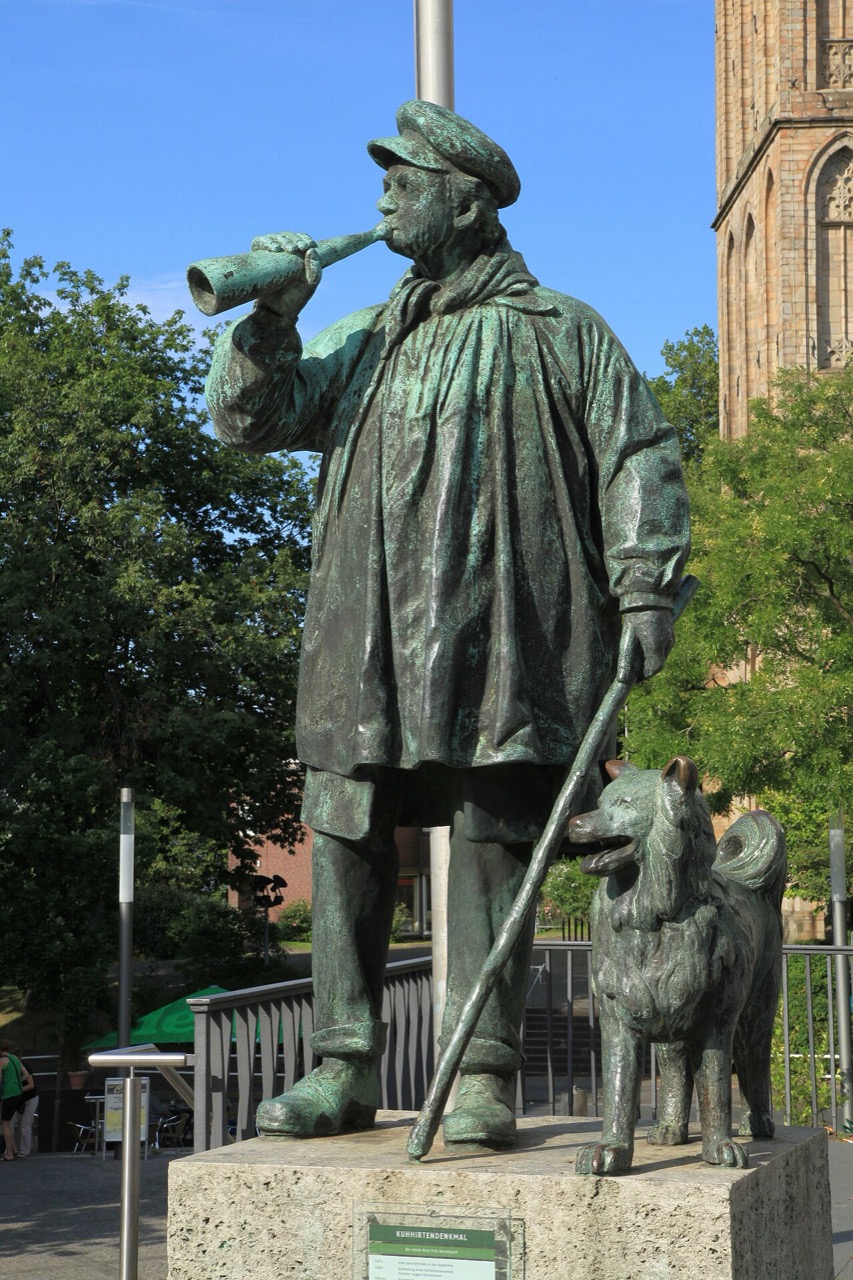 Eine grüne Bronzefigur eines Mannes mit Kappe und langem Umhang, der in ein Horn bläst. In der anderen Hand hält er einen langen Stab. Neben ihm steht ein Hund. Die Figur steht auf einem niedrigen Sockel aus grauem Stein mit einer kleinen Tafel. Dahinter liegen ein Geländer, Bäume und ein Kirchturm aus Sandstein.