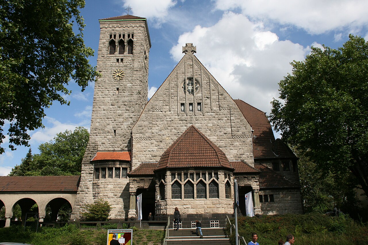 Eine Kirche aus grauem Naturstein. Links steht ein hoher, schlanker Turm mit vier schmalen Schallöffnungen am oberen Ende. In der Mitte ragt ein steiler Giebel mit rundem Fenster auf. Davor tritt ein Vorbau mit vieleckigem, braunrotem Dach und Spitzbogenfenstern hervor. Links schließt ein Gang mit Rundbögen und rotem Ziegeldach an. Bäume stehen zu beiden Seiten.