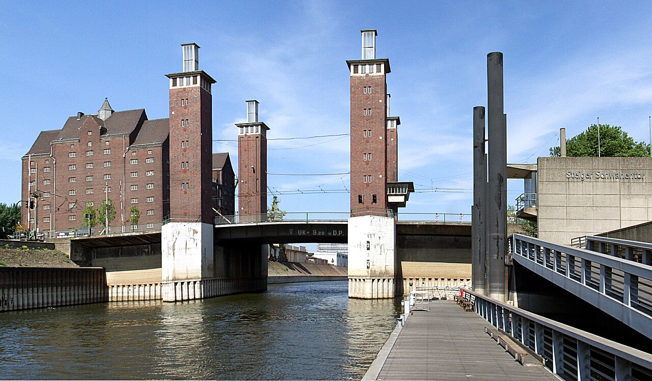 Eine Brücke über ein Hafenbecken. Vier schlanke Türme aus rotbraunem Backstein stehen paarweise zu beiden Seiten der Fahrbahn, ihre Sockel sind aus hellem Beton. Zwischen den Türmen liegt eine flache Brückenplatte. Links dahinter steht ein langes, mehrgeschossiges Backsteinhaus mit Fensterreihen. Rechts führt ein Holzsteg mit Metallgeländer am Wasser entlang.