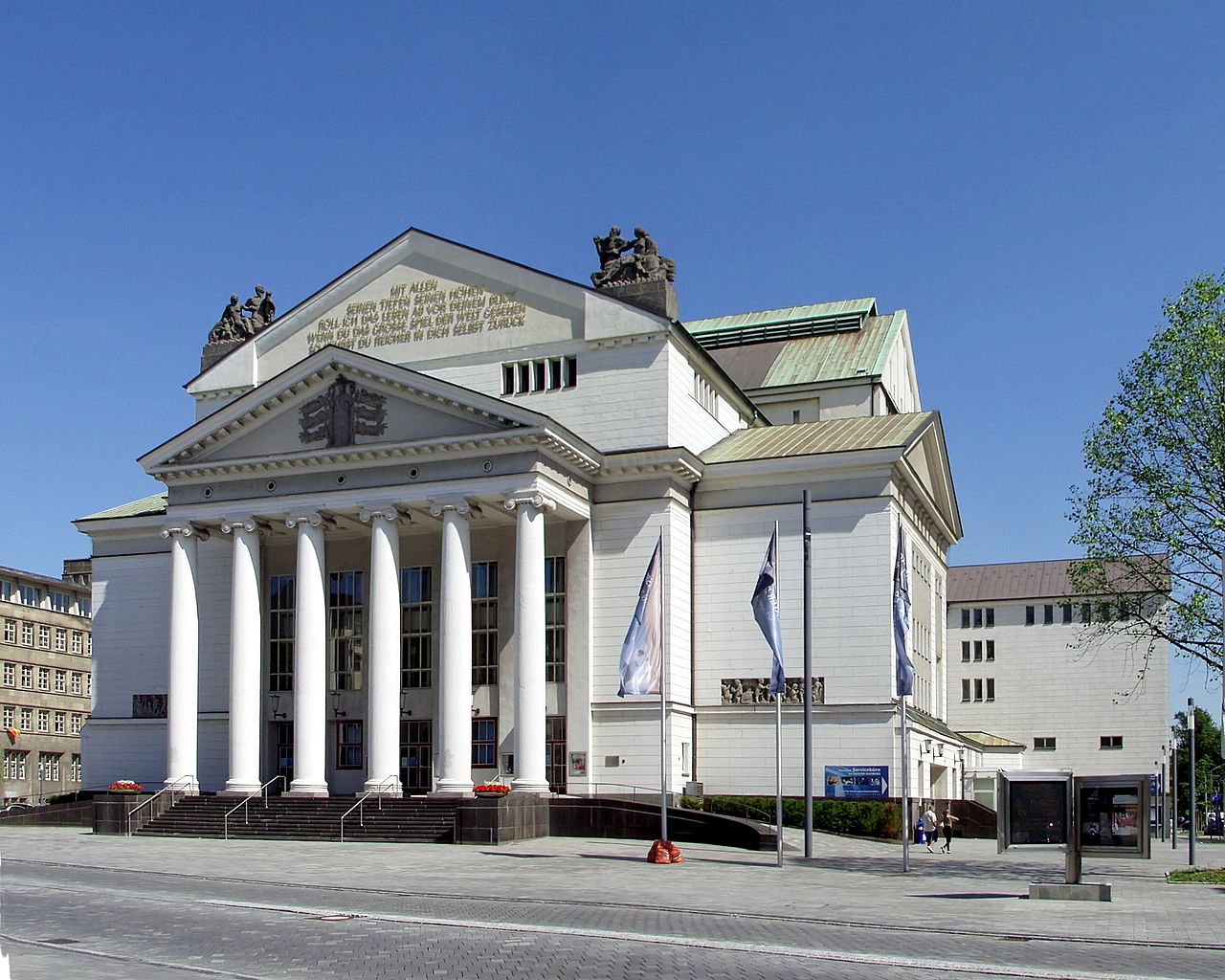 Ein weißer Bau mit einer Säulenhalle aus sechs hohen Säulen. Über der Halle liegt ein Dreiecksgiebel mit Wappenrelief. Dahinter ragt ein höherer Bauteil mit einer Inschrift im Giebel auf, auf dessen Ecken Figurengruppen stehen. Das Dach ist mit grünem Metall gedeckt. Rechts schließen niedrigere, helle Bauteile an. Davor liegt ein gepflasterter Vorplatz.
