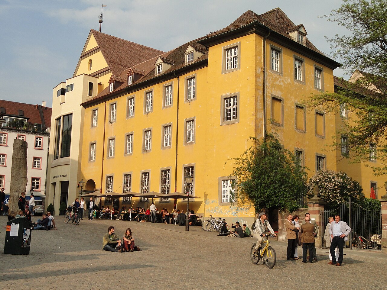 Ein langgestrecktes Gebäude mit ockergelber Fassade und dunklem Ziegeldach mit Gauben. Die hohen Fenster sind in regelmäßigen Reihen angeordnet und hell gerahmt. Links schließt ein moderner Bau mit großen Glasflächen an. Davor liegt ein weiter, gepflasterter Platz, rechts steht ein Laubbaum.