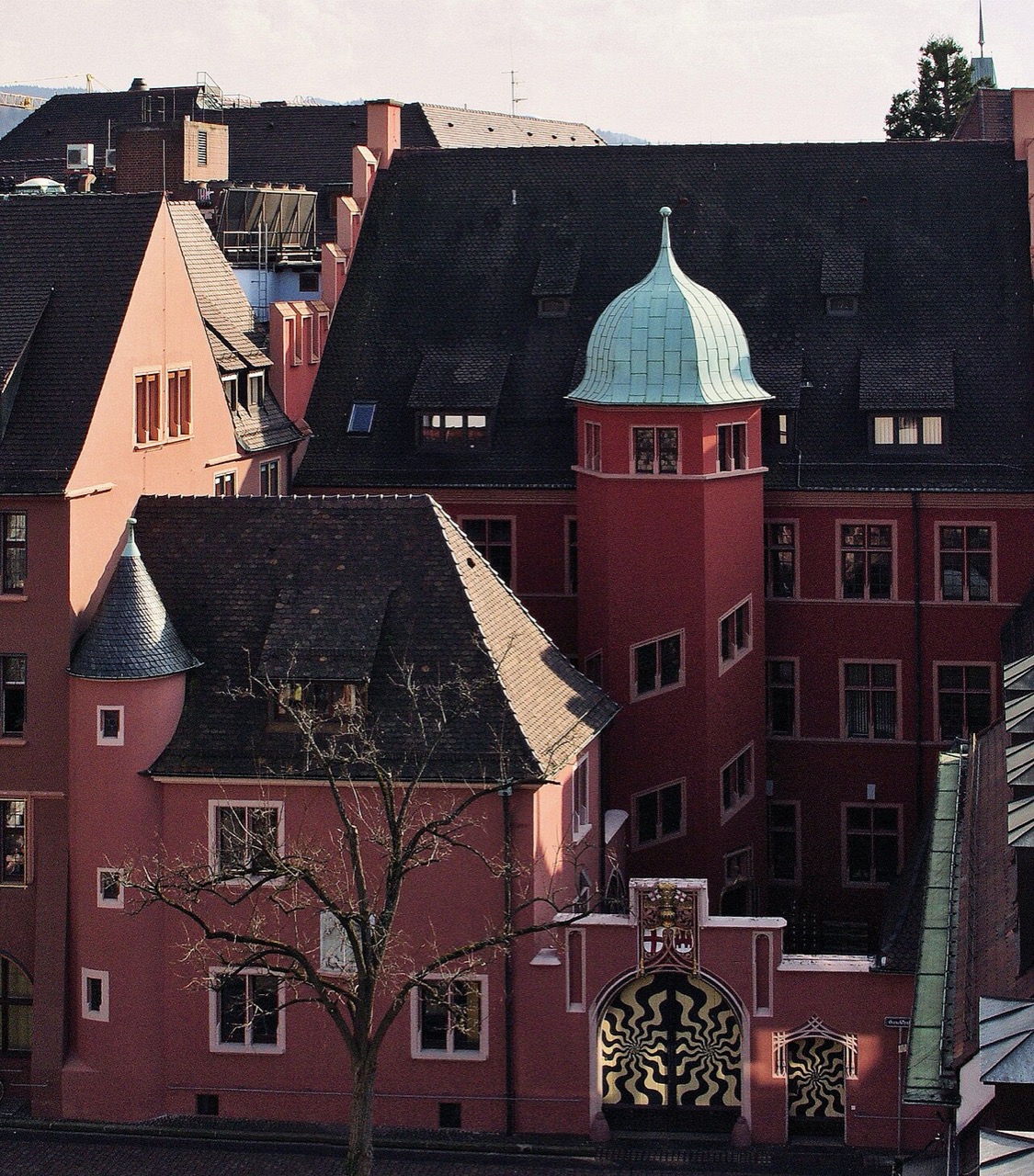 Ein großes, rosarot verputztes Haus mit steilem, dunklem Dach. An der Ecke steht ein runder Treppenturm mit grüner Kupferhaube und Spitze. Die Fenster sind klein und weiß gerahmt. Links schließt ein ebenfalls rosafarbener Bau mit kleinem, rundem Eckturm an. Unten sitzt ein Portal mit goldfarbenen Zierformen vor schwarzem Gitterwerk.