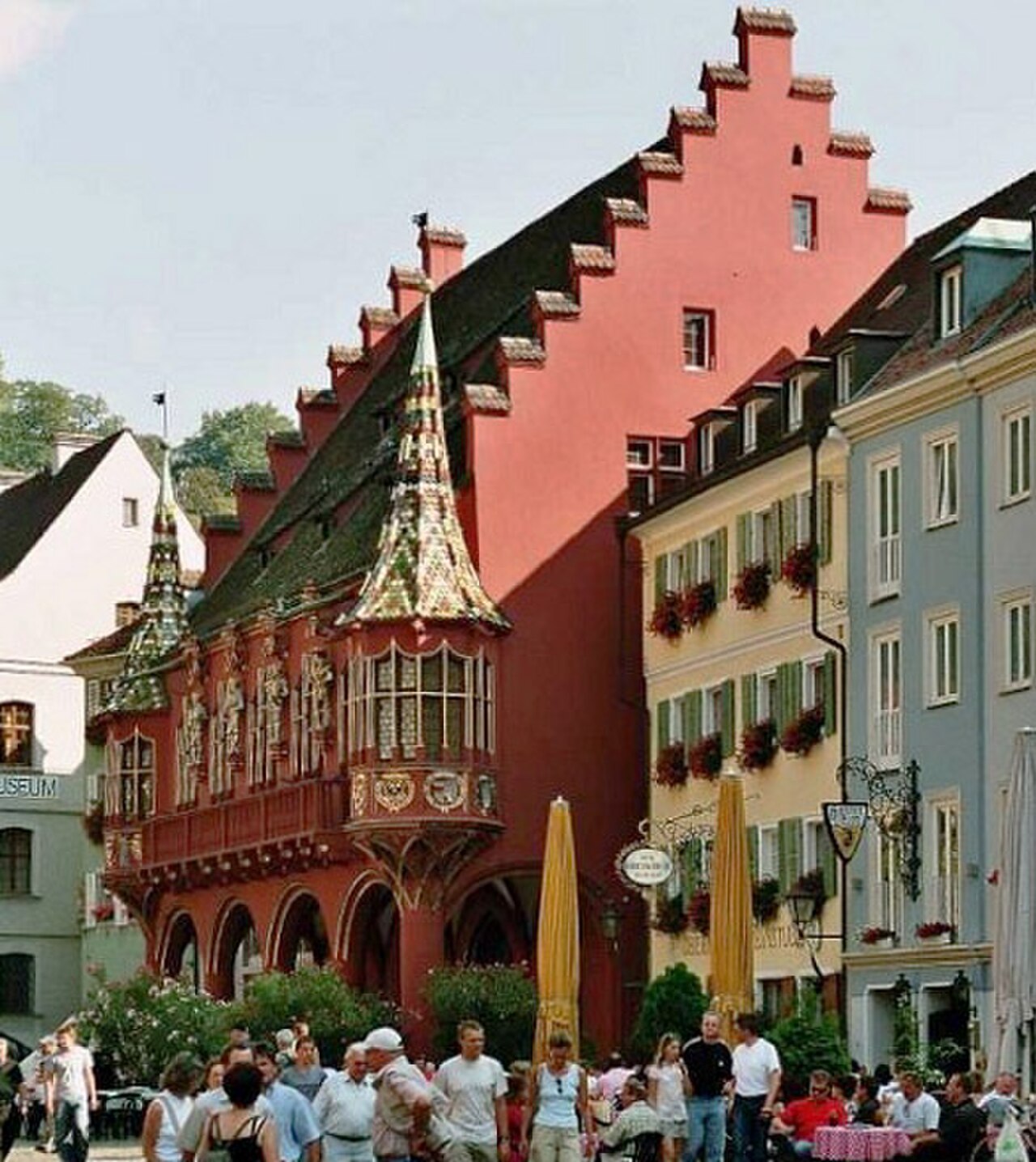 Ein rotes Gebäude mit steilem Dach und treppenförmig abgestuftem Giebel. Im Erdgeschoss öffnen sich Rundbögen, darüber läuft ein Balkon. An der Ecke sitzt ein zweigeschossiger Erker mit schlanker, bunt gedeckter Spitze. Rechts stehen gelbe und blaugraue Bürgerhäuser mit grünen Fensterläden.