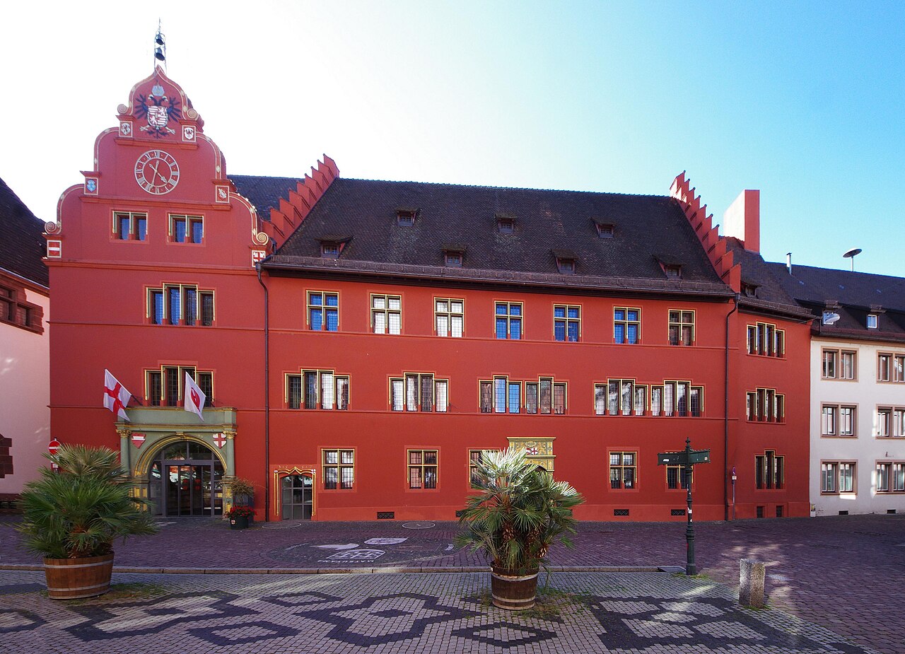 Ein langgestrecktes, rot gestrichenes Gebäude mit dunklem Dach und Gauben. Links ragt ein schmaler, ebenfalls roter Giebelturm mit Uhr und kleiner, grüner Haube auf. An den Dachenden steigen Treppengiebel an. Unten links öffnet sich ein Rundbogenportal unter einem grünen Vorbau. Davor liegt ein Platz mit hellem und dunklem Steinmuster.