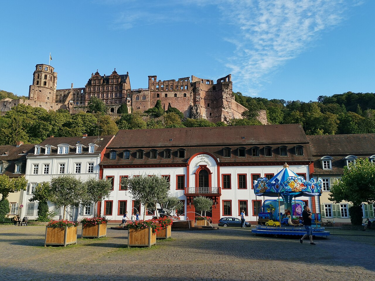 Ein langgestrecktes Haus mit hellem Putz und flachem, dunkelrotem Dach mit Gauben. In der Mitte springt ein Bauteil mit rotem Rahmen und Rundbogen vor, dort liegt der Eingang. Links steht ein weißes Haus mit regelmäßigen Fensterreihen. Davor liegt ein Platz mit Pflanzkästen. Hinter den Dächern steigt ein bewaldeter Hang mit einer Schlossruine auf.