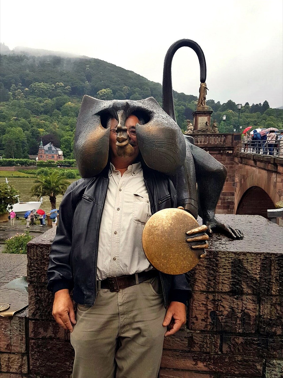 Eine Bronzefigur eines Affen sitzt auf einer Brüstung aus rotem Sandstein. Der Kopf ist groß, mit Ohren. In der Hand hält die Figur eine große, runde Scheibe aus goldfarbenem Metall. Hinter ihr ragt ein gebogener Stab auf. Rechts führt die Brücke mit gemauerten Bögen zum anderen Ufer. Dahinter steigt ein bewaldeter Hang an.