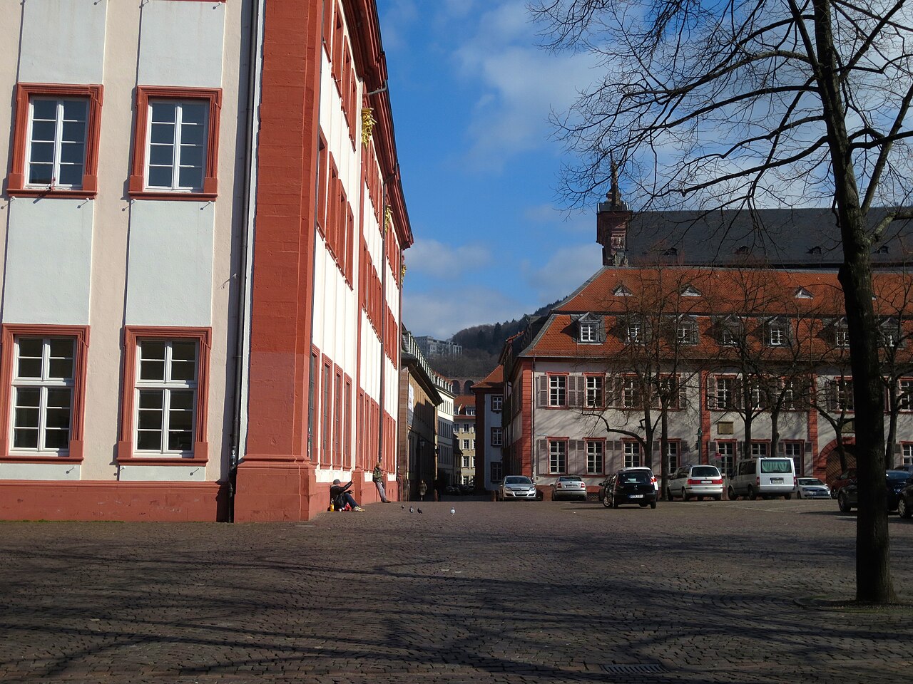 Ein weiter Platz mit Kopfsteinpflaster. Links steht ein langes, mehrgeschossiges Gebäude mit hellem Putz. Ecken und Rahmen der Fenster bestehen aus rotem Sandstein. Rechts im Hintergrund steht ein Haus mit rotem Ziegeldach, daneben ein Turm mit dunklem Dach. Ein Baum steht rechts im Vordergrund.