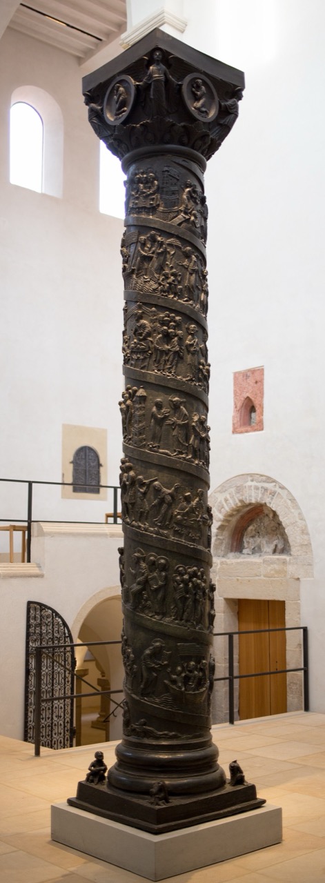 Eine hohe, dunkle Bronzesäule steht in einem hellen Raum mit weiß verputzten Wänden. Ihr Schaft ist in übereinanderliegenden Bändern mit Reliefs bedeckt, dicht gedrängten Szenen kleiner Figuren. Oben trägt sie ein ausladendes Kapitell mit runden Medaillons, unten einen hellen Steinsockel, an dessen Ecken kleine Figuren hocken. Rechts liegt eine Holztür unter einem steinernen Rundbogen.