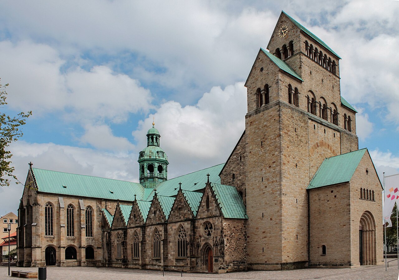 Eine große romanische Kirche aus hellbraunem Sandstein mit grün patinierten Kupferdächern. Rechts steht ein breiter, hoher Turm mit steilem Dach und Rundbogenfenstern. Das lange Schiff links trägt eine Reihe spitzer Giebel mit Spitzbogenfenstern. Aus dem Dach ragt ein kleiner Turm mit grüner Kuppel.