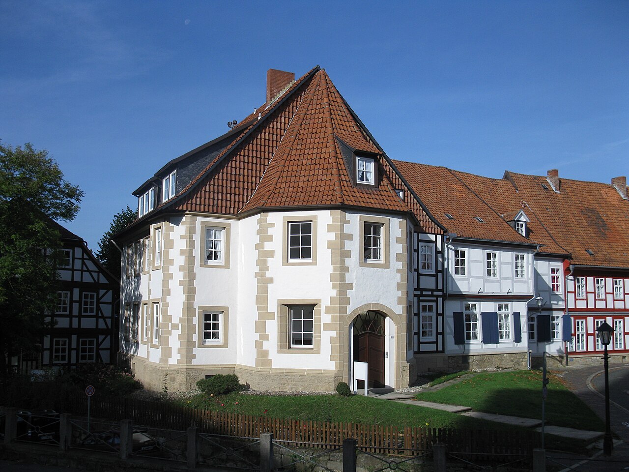 Ein kompaktes Haus mit weiß verputzten Wänden und vieleckig vorspringender Front. Kanten und Fensterrahmen sind aus hellem Sandstein, an der Seite sitzt ein Rundbogenportal mit Holztür. Das Dach ist steil, mit roten Ziegeln gedeckt und läuft an der Front spitz zusammen. Rechts schließen Fachwerkhäuser an, davor liegen eine Wiese und ein niedriger Holzzaun.
