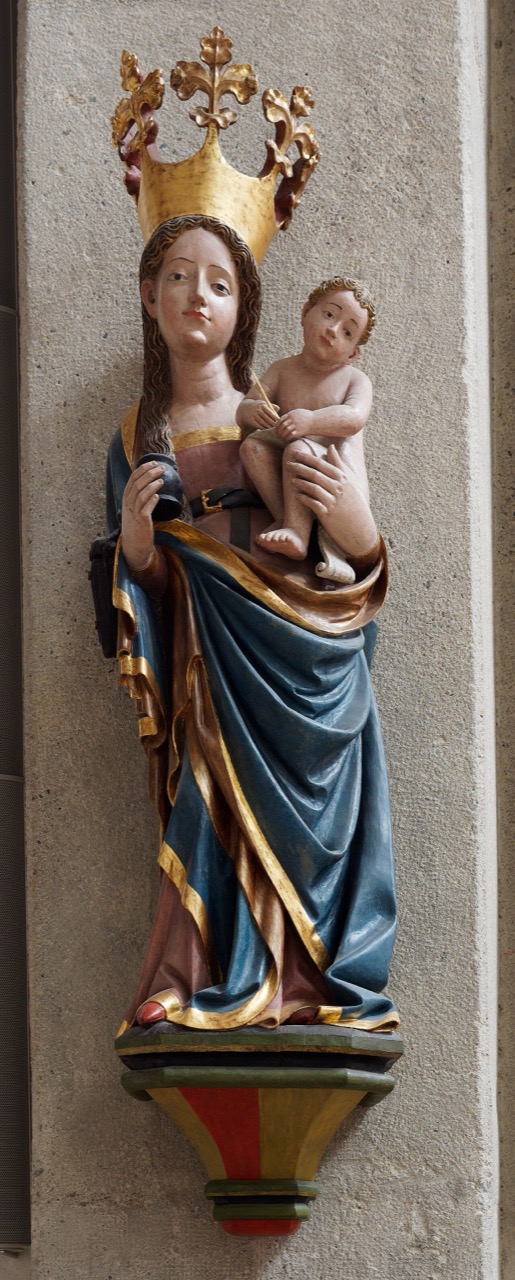Eine farbig gefasste, aufrechte Marienfigur steht auf einer kleinen Konsole an einem hellen Steinpfeiler. Sie trägt eine goldene, zackige Krone, langes, welliges Haar und einen blauen Mantel mit goldenem Saum. Auf dem linken Arm sitzt das nackte Kind mit lockigem Haar und hält einen dünnen Stift. In ihrer Hand liegt ein kleiner, dunkler Gegenstand.