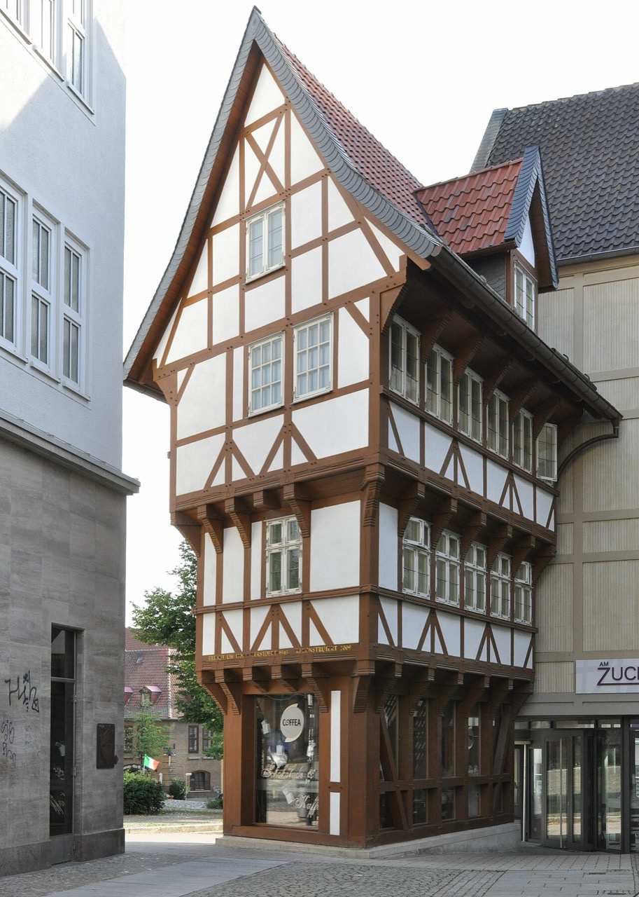 Ein schmales Fachwerkhaus, das nach oben breiter wird: Jedes Geschoss ragt weiter vor als das untere. Die Balken sind braun, die Gefache weiß, das Dach ist steil und rot gedeckt. Im Erdgeschoss liegt ein Schaufenster. Rechts schließt ein größeres Haus mit grauem Dach an, links steht ein heller Putzbau.