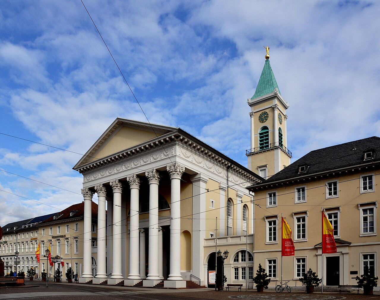 Eine klassizistische Kirche mit Säulenvorhalle. Hohe, weiße Säulen mit verzierten Kapitellen tragen ein Gebälk und einen flachen Dreiecksgiebel in hellem Gelb. Dahinter steht ein viereckiger Turm mit Uhr und grünem Kupferhelm. Die Wände sind gelblich verputzt. Rechts schließt ein langes Haus mit dunklem Dach an, links eine lange Zeile heller Häuser.