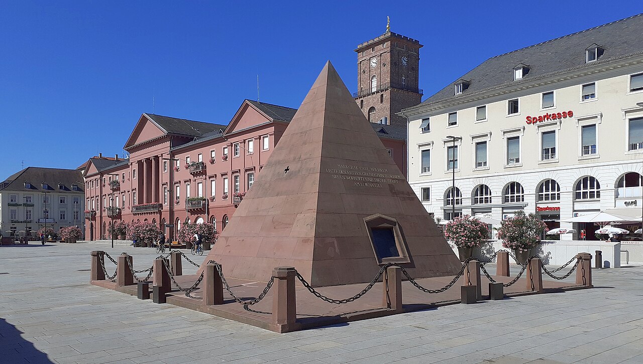 Eine Pyramide aus rotbraunem Sandstein mit glatten, steil ansteigenden Flächen. An einer Seite sitzt eine kleine, dunkle Öffnung mit Rahmen. Um den Sockel steht ein niedriger Zaun aus Steinpfosten, die durch dunkle Ketten verbunden sind. Sie steht auf einem weiten, hell gepflasterten Platz. Dahinter erkennt man ein rosafarbenes Gebäude mit Turm, rechts ein langes, helles Haus.