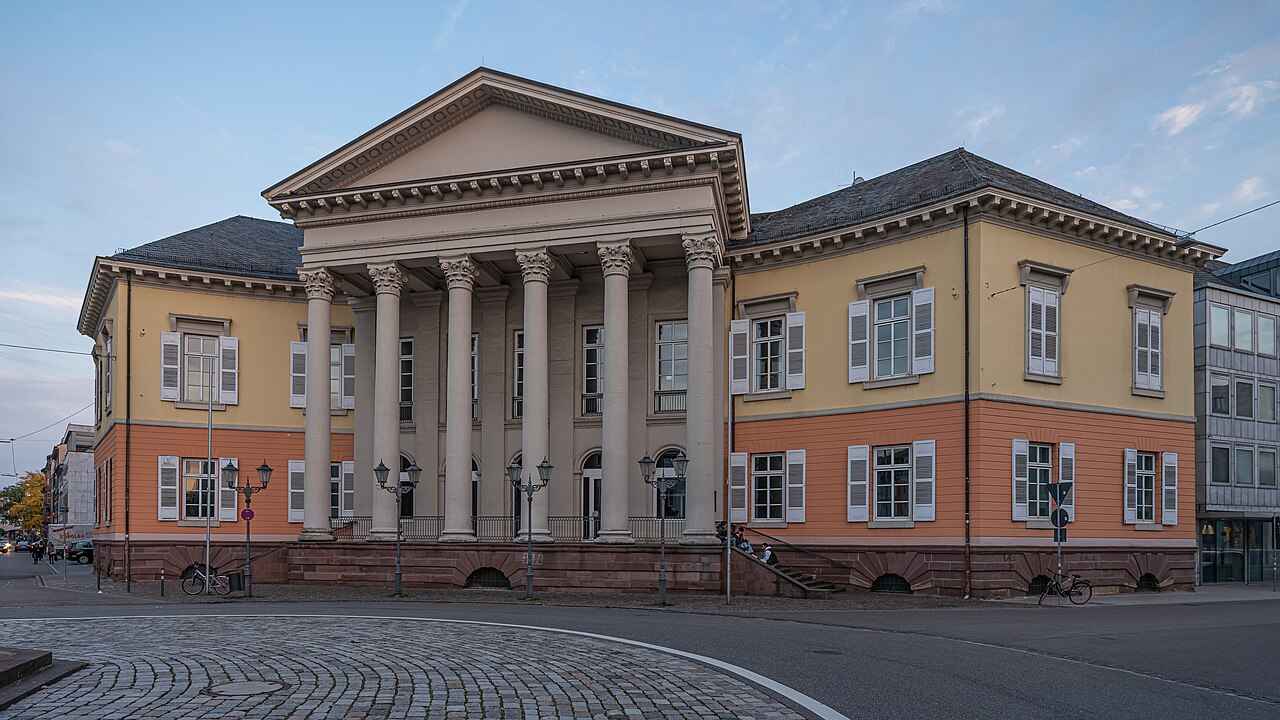 Ein breites, klassizistisches Gebäude mit einem Säulenportikus in der Mitte. Sechs hohe Säulen tragen einen flachen Dreiecksgiebel. Die beiden Seitenflügel sind oben gelb, unten orange verputzt, mit weißen Fensterläden und dunklen Dächern. Der Sockel besteht aus rotem Sandstein. Davor liegt ein gepflasterter Platz mit Straßenlaternen.