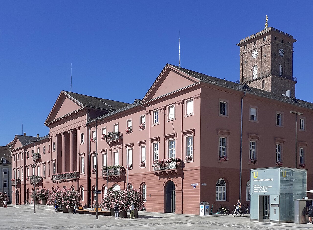 Ein langgestrecktes, rosafarbenes Gebäude mit klassizistischer Gliederung auf einem Sockel aus rotem Sandstein. Zwei flache Dreiecksgiebel setzen Akzente, links öffnet sich eine Säulenhalle. Kleine Balkone mit Gittern und Rundbogentüren im Erdgeschoss. Hinter dem Dach ragt ein viereckiger Turm aus braunrotem Stein mit Uhr und goldenem Kreuz auf. Rechts steht ein kleiner Glasbau mit U-Zeichen.