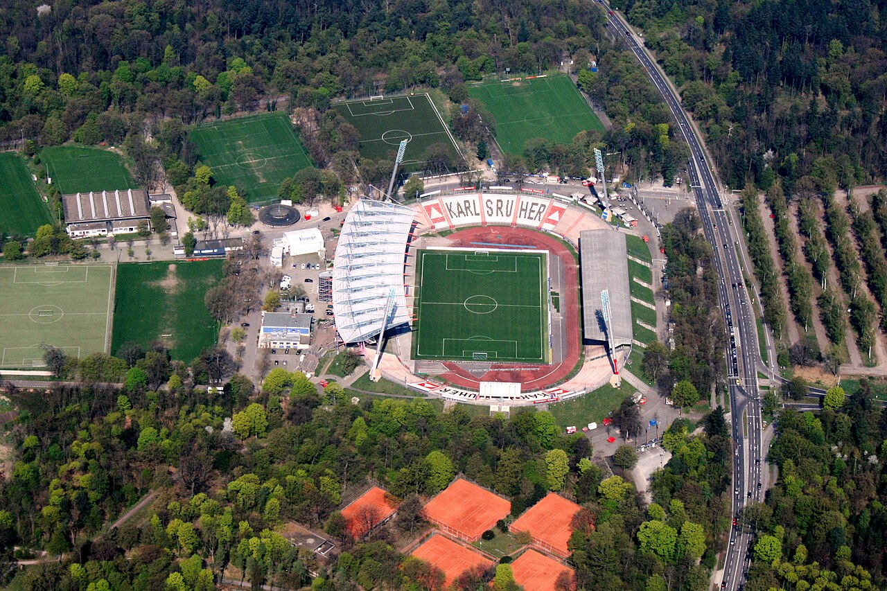 Ein ovales Stadion mit grünem Spielfeld, umgeben von einer roten Laufbahn. Links überspannt ein weißes, geschwungenes Dach die Ränge, rechts steht ein flacheres, graues Dach. Das Stadion liegt im Wald. Daneben liegen mehrere Rasenplätze, unterhalb rote Tennisplätze. Rechts verläuft eine lange, gerade Straße.
