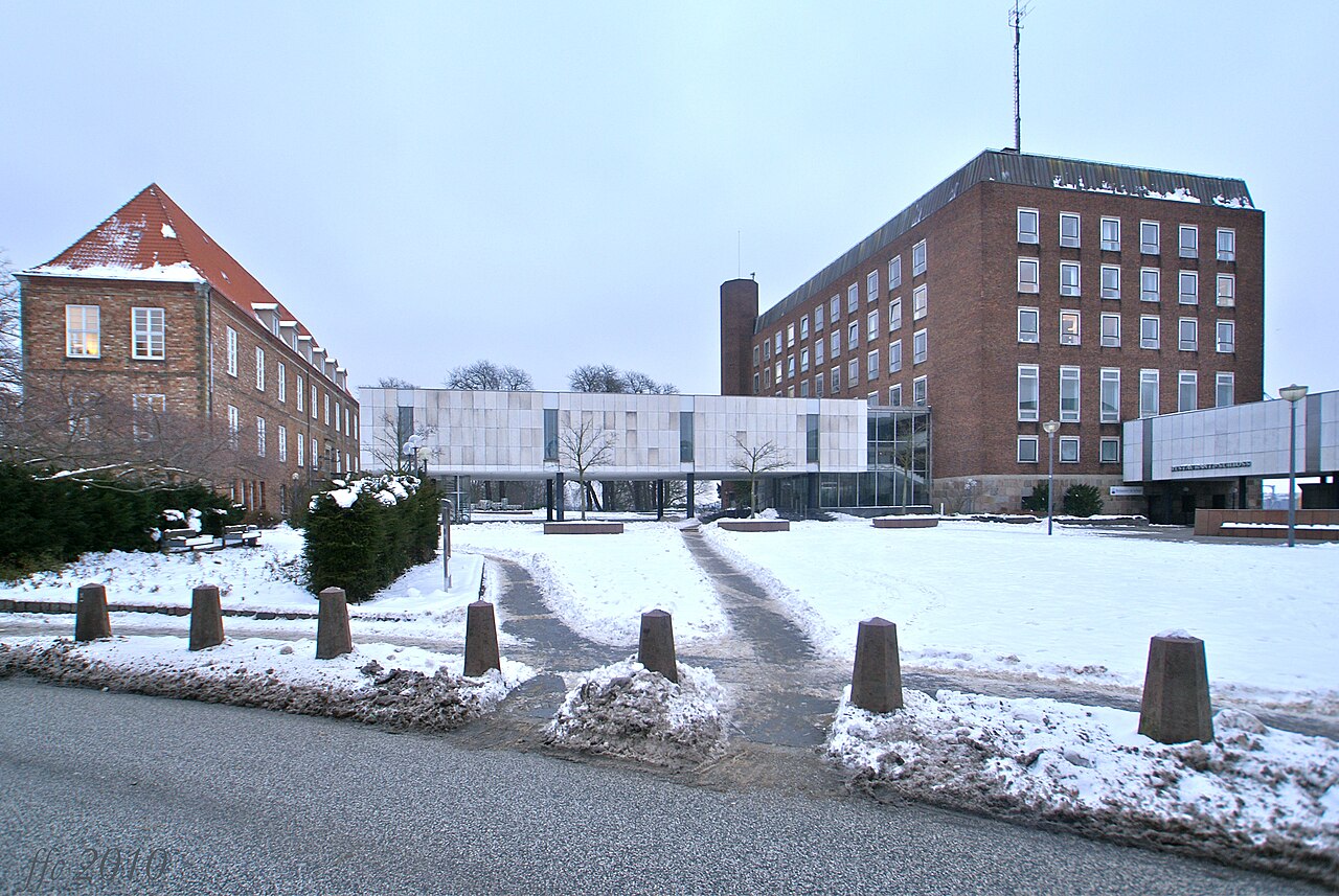 Mehrere Backsteinbauten um einen weiten Platz. Rechts steht ein hoher, rotbrauner Block mit regelmäßigen Fensterreihen und einem runden Schornstein an der Ecke. Links liegt ein Haus mit steilem, rot gedecktem Dach und Giebel. Dazwischen steht ein flacher Bau mit Glasfront und hellen Stützen. Davor reihen sich niedrige Steinpoller.