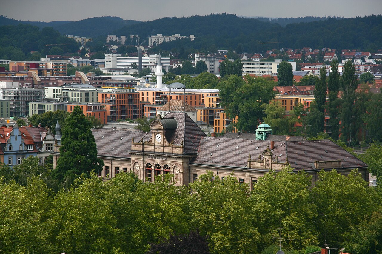 Ein langgestrecktes Schulhaus aus hellbraunem Stein mit flachem, grauem Schieferdach. In der Mitte sitzt über einer Reihe von Rundbogenfenstern ein Giebel mit runder Uhr. Rechts auf dem Dach steht ein kleines Türmchen mit grüner Kupferhaube. Hohe Laubbäume stehen davor. Dahinter liegen neuere Wohn- und Bürohäuser, am Horizont ein bewaldeter Höhenzug.