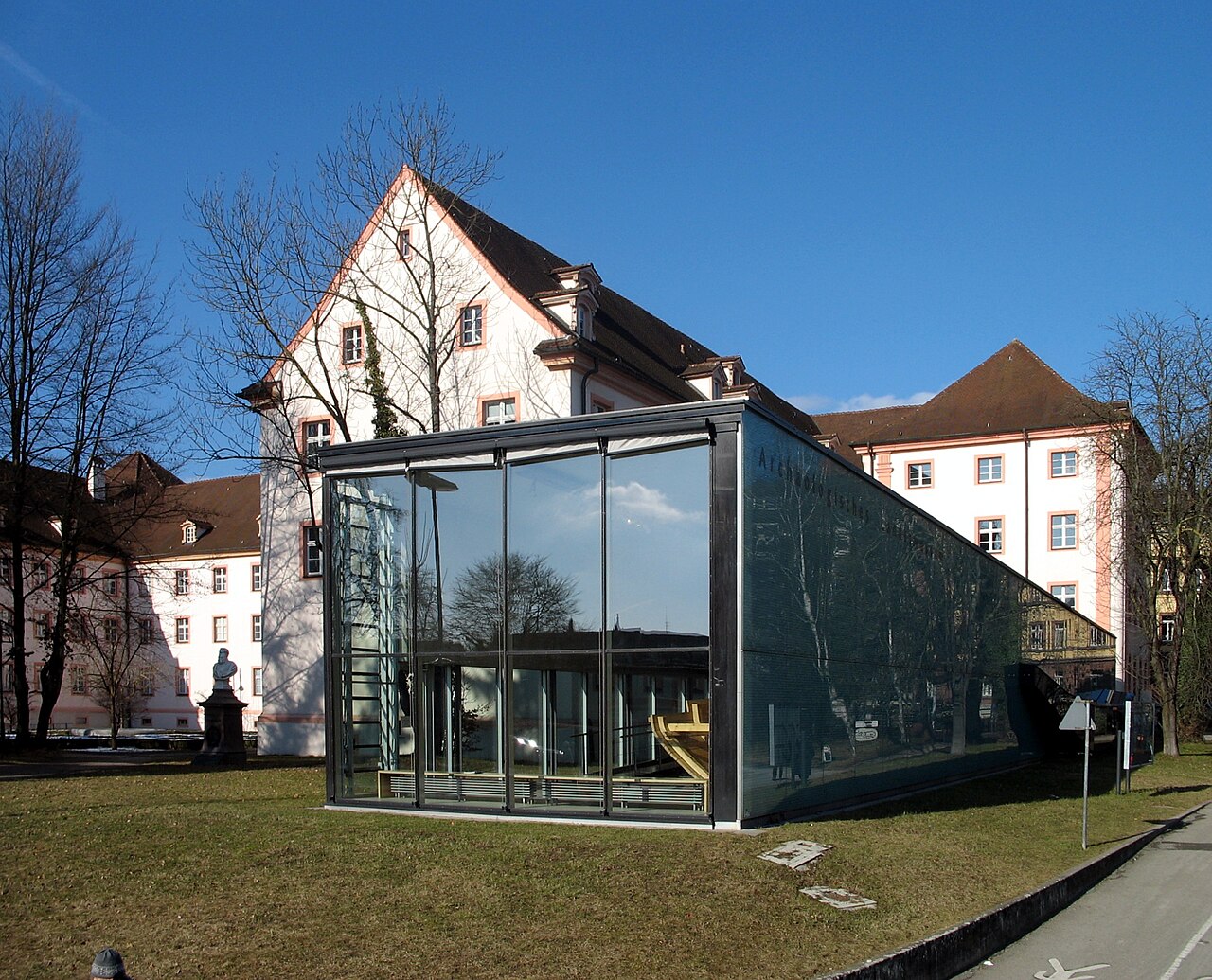 Ein moderner Glasbau mit schmalem, dunklem Rahmen steht auf einer Rasenfläche. Die Vorderseite ist senkrecht verglast, die Seitenwand fällt schräg zum Boden ab. Im Inneren ist ein gelblicher Gegenstand zu erkennen. Dahinter steht ein hell verputztes, mehrgeschossiges Haus mit steilem Satteldach und rosa gerahmten Fenstern, rechts ein zweites mit Walmdach.