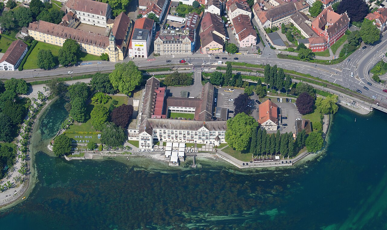 Eine kleine Landzunge ragt ins Wasser. Auf ihr steht ein Gebäudekomplex mit hellen Wänden und grau-braunen Dächern, die lange Hauptfront mit Fensterreihen liegt am Ufer. Rechts steht ein kleineres Haus mit orangerotem Dach, davor wachsen Bäume. Links liegen Gartenflächen mit Wiese. Hinter der Landzunge verläuft eine breite Straße, dahinter stehen Wohn- und Geschäftshäuser.