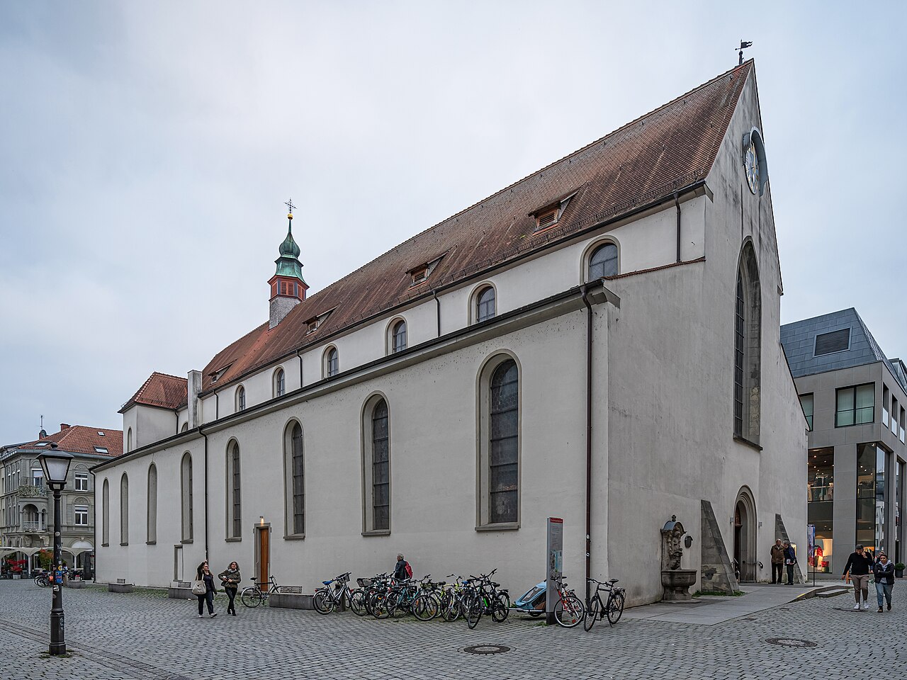 Eine lange, schlichte Kirche mit hellgrau verputzten Wänden und steilem, rotbraunem Ziegeldach. Hohe Spitzbogenfenster reihen sich an der Längsseite, darüber sitzen kleine Dachfenster. Am linken Ende ragt ein Dachreiter mit grüner Haube auf. Die rechte Giebelseite trägt ein sehr hohes, schmales Spitzbogenfenster. Davor liegt ein gepflasterter Platz.