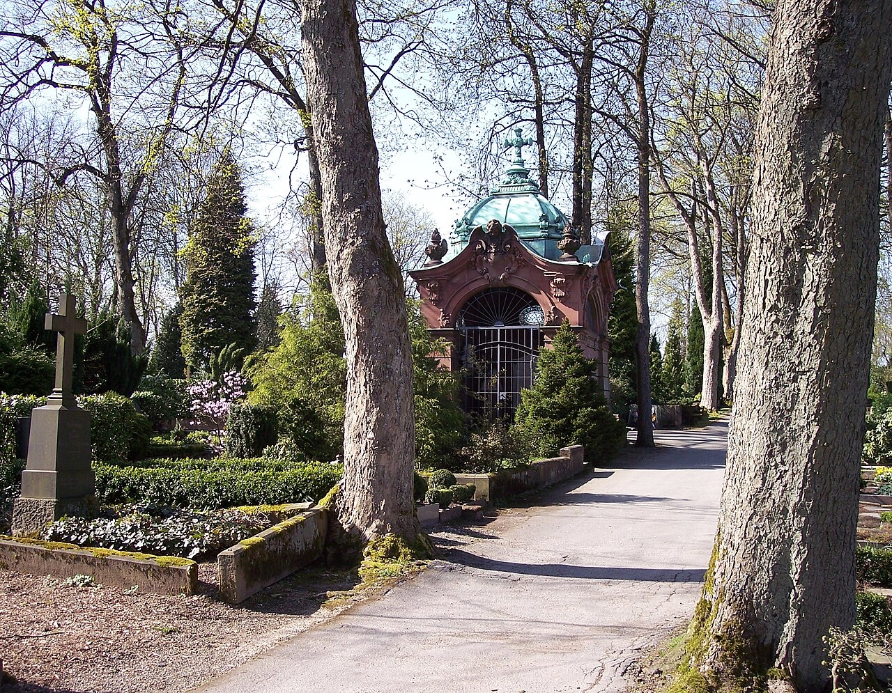 Ein kleines Mausoleum aus rotbraunem Stein mit einem grünen Kupferdach in Kuppelform, das ein Kreuz trägt. Die Vorderseite öffnet sich in Rundbögen, die mit Eisengittern geschlossen sind. Es steht hinter Hecken und Nadelgehölzen an einem schmalen, hellen Weg. Im Vordergrund ragen links und rechts dicke Baumstämme auf, links ein hoher grauer Grabstein.