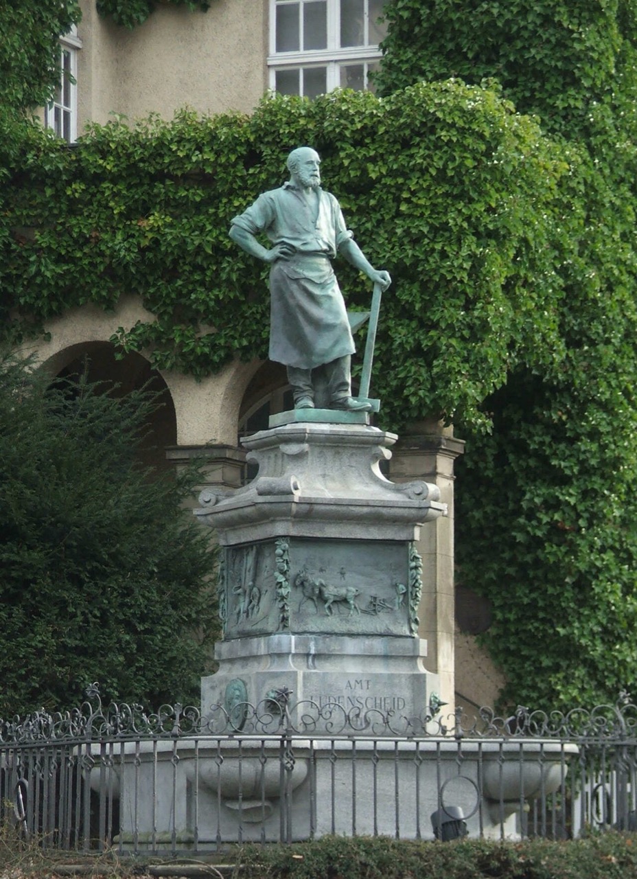Ein Standbild aus grünlicher Bronze: ein bärtiger Mann in langer Schürze stützt sich mit einer Hand auf ein langes Werkzeug. Er steht auf einem hohen, gegliederten Steinsockel mit einer Bildplatte und einer Inschrift. Unten umgibt ein schwarzes Eisengitter ein flaches Brunnenbecken. Dahinter ist die Wand eines Hauses dicht mit Efeu bewachsen.