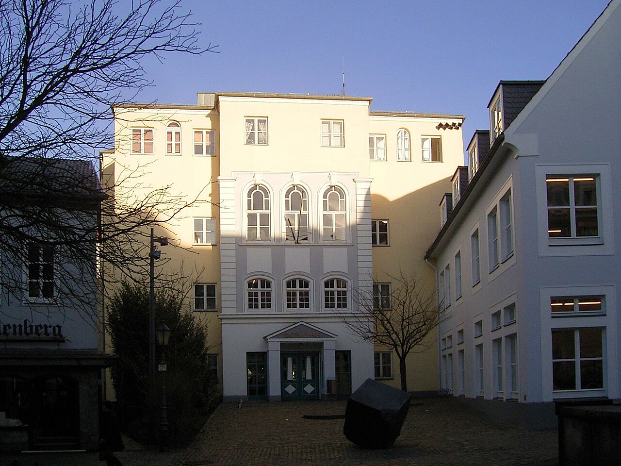 Ein hohes, gelb verputztes Gebäude. Der Mittelteil tritt vor, oben sitzen drei Rundbogenfenster, darunter zwei kleinere Fenster und ein Eingang mit Glastür unter einem kleinen Vordach. Die Seitenteile haben schlichte, kleine Fenster. Rechts schließt ein weißes Haus mit dunklem Dach und weiß gerahmten Fenstern an. Davor liegt ein Platz mit rotbraunem Pflaster.