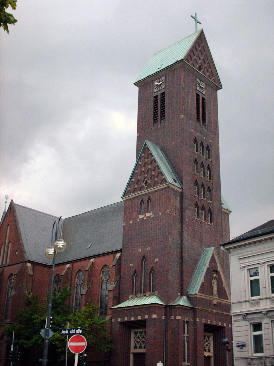 Eine Backsteinkirche in dunklem Rot. Der hohe, viereckige Turm trägt ein grünes Kupferdach in Pyramidenform mit Kreuz, an den Seiten sitzen Zifferblätter. In den Turm sind schmale, hohe Bogenfenster eingelassen, davor stehen Ziergiebel mit grünen Dachkanten. Links schließt das Kirchenschiff mit grauem Schieferdach an. Davor steht eine Straßenlaterne, rechts ein helles Haus.