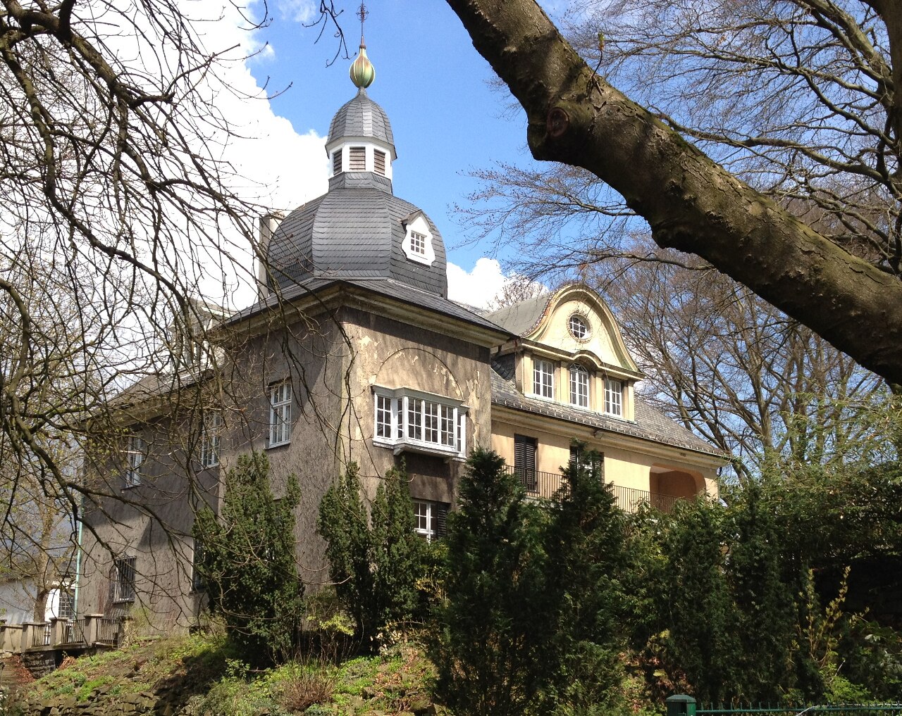 Eine Villa aus graubraunem Stein in einer begrünten Anlage. Über dem Mittelteil sitzt ein rundes, grau gedecktes Kuppeldach mit kleiner Laterne und goldener Kugel an der Spitze. Das Haus hat ein Erkerfenster mit weiß gerahmten Sprossenfenstern. Rechts schließt ein Flügel mit gelbem Giebel und rundem Fenster an. Davor stehen dunkelgrüne Nadelgehölze.