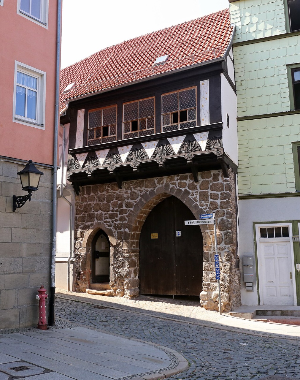 Ein schmales Haus mit massivem Erdgeschoss aus grauem Bruchstein. Darin öffnet sich ein großes Spitzbogentor mit dunklen Holztüren, links daneben ein kleineres Spitzbogenportal. Das Obergeschoss kragt vor und besteht aus dunklem Fachwerk mit hellen Feldern und Fenstern mit Sprossengittern. Unter dem Überstand sitzen Schnitzereien in Fächerform. Das Dach ist steil und rot gedeckt.