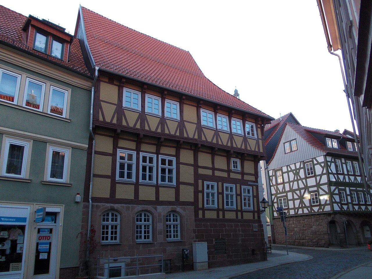Ein mehrgeschossiges Fachwerkhaus mit dunkelbraunen Balken und gelb-ockerfarbenen Gefachen unter einem steilen, roten Dach. Das Erdgeschoss ist massiv und hat drei Rundbogenfenster mit Gittern. Darüber sitzen Reihen kleiner Fenster. Links steht ein heller Putzbau mit einem Geschäft im Erdgeschoss, rechts folgt ein weiteres Fachwerkhaus.