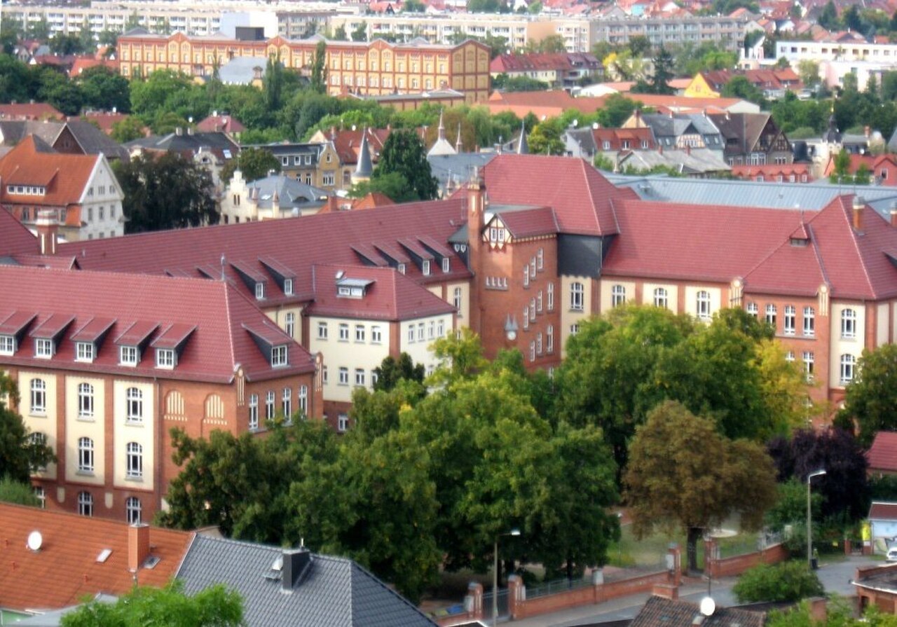 Ein großes Schulgebäude mit mehreren Flügeln unter steilen, rotbraunen Ziegeldächern mit zahlreichen Gauben. Die Fassaden sind hell verputzt, teils rötlich, mit gleichmäßigen Fensterreihen. Große Laubbäume stehen vor den Flügeln. Dahinter liegen weitere Gebäude mit Satteldächern, in der Ferne ein langer Bau mit orangem Dach.