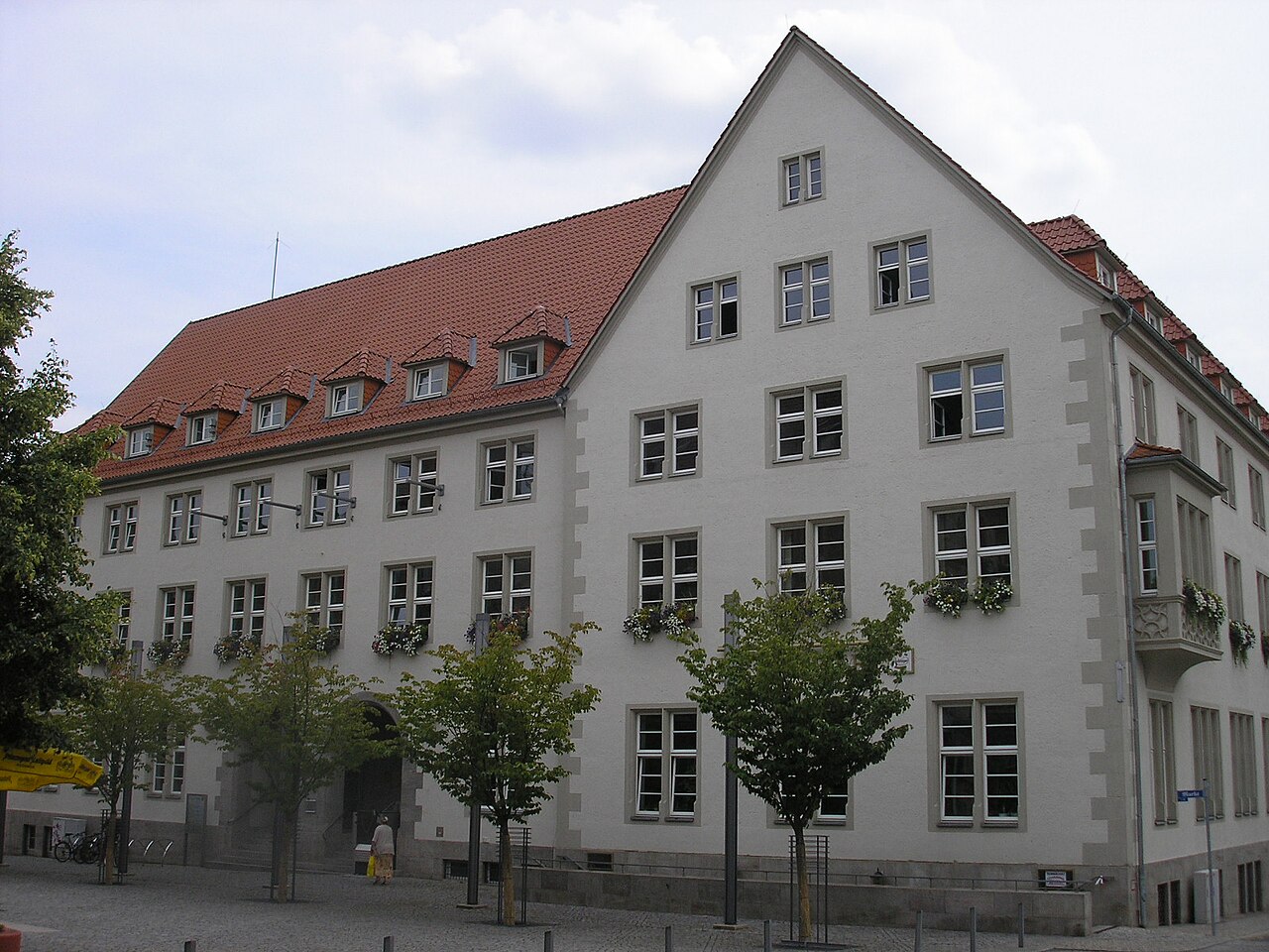 Ein großes, hell verputztes Haus mit steilem, rotem Ziegeldach. Links erstreckt sich ein langer Flügel mit zwei Reihen kleiner Dachgauben, rechts steht ein Querbau mit hohem Giebel. Die Fenster stehen in gleichmäßigen Reihen, an manchen hängen Blumenkästen. Davor stehen junge Bäume.