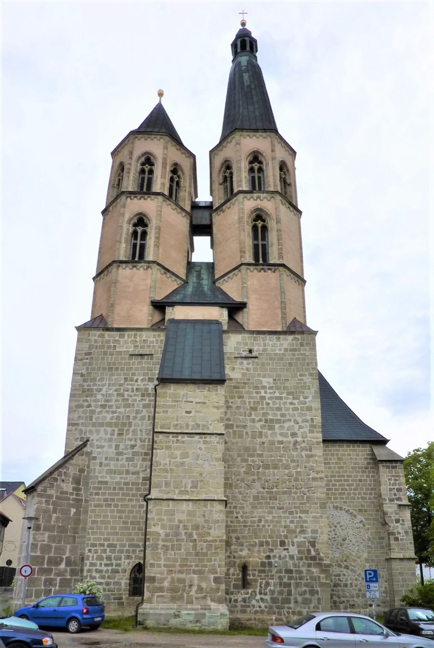 Eine Kirche mit zwei Türmen über einem breiten, massiven Unterbau aus grauem Bruchstein. Die Türme sind aus rosafarbenem Stein, haben gekoppelte Rundbogenfenster und enden in dunklen, spitzen Dächern. Der rechte Turm ist höher und trägt eine schlanke Spitze. Ein kleines Zwischenstück verbindet beide Türme.