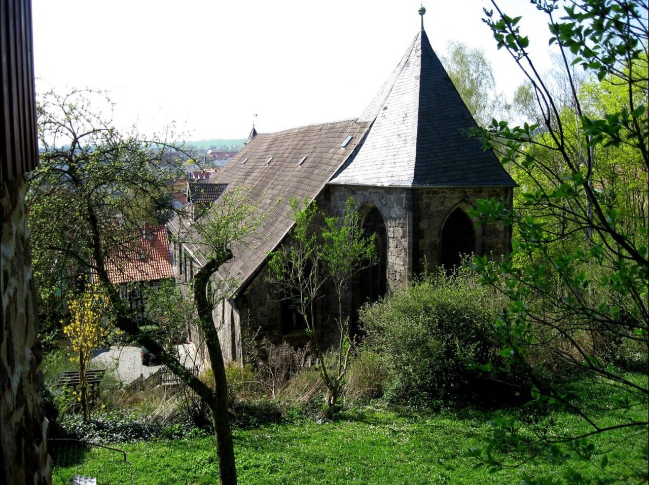 Ein kleines Steingebäude mit hohem, spitzem Dach aus dunklem Schiefer. Es ist aus grauem Bruchstein gemauert, in der Wand sitzen hohe Rundbogenöffnungen. An das spitze Dach schließt ein niedrigeres, langes Satteldach an. Das Gebäude steht in einer Wiese, Sträucher und Bäume verdecken Teile davon.