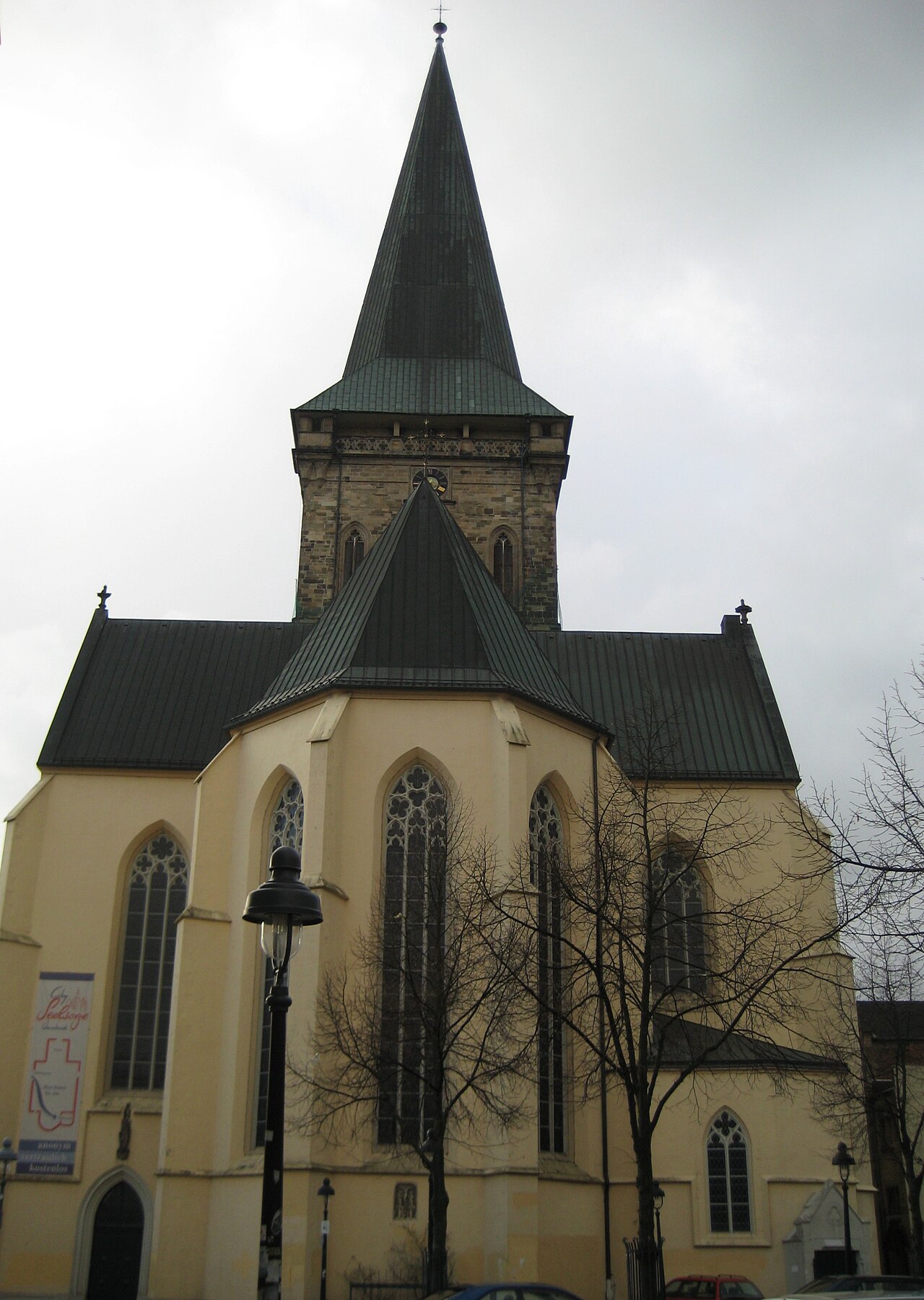 Eine große Kirche mit gelb verputztem, vieleckigem Chor und dunkelgrünen Metalldächern. Hohe Spitzbogenfenster mit Maßwerk gliedern den Chor. Dahinter erhebt sich ein Turm aus Naturstein mit Uhr und einer sehr hohen, schlanken, dunkelgrünen Spitze. Links unten liegt ein Spitzbogenportal. Davor stehen eine Laterne und kahle Bäume.