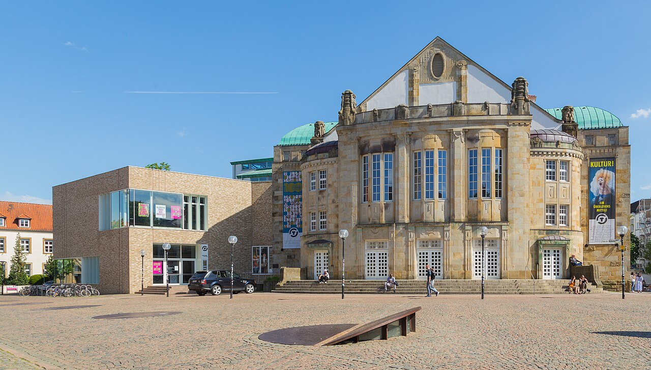 Rechts steht ein Bau aus gelbem Sandstein mit hohem Giebel, in dem eine ovale Öffnung sitzt. Gerundete Seitenteile tragen grüne Kupferkuppeln, auf dem Gesims sitzen steinerne Figuren. Hohe Fenster stehen in Dreiergruppen, unten führen flache Stufen zu weißen Gittertüren. Links steht ein moderner Bau aus braunem Backstein mit breitem Glasband. Davor liegt ein weiter Platz aus Kopfsteinpflaster.