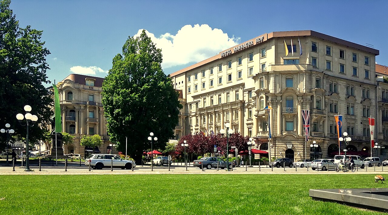 Ein großes, mehrgeschossiges Hotel an einer Straßenecke mit heller, gelblicher Fassade. Die Fenster stehen in dichten Reihen, im Erdgeschoss sitzen Rundbögen. Die Front hat Balkone und Fahnenmasten. Davor liegen eine Rasenfläche, eine Straße und hohe Bäume. Links daneben steht ein kleineres Haus.