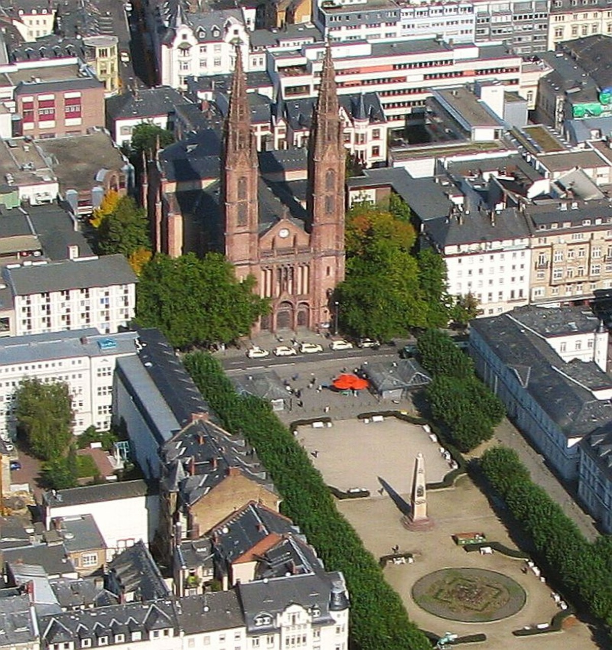 Ein großer, rechteckiger Platz mit heller Kiesfläche. In der Mitte steht ein Obelisk, daneben liegt ein rundes Beet mit Muster. An einer Seite verläuft eine Reihe Bäume. An einem Ende des Platzes steht eine rote Kirche mit zwei hohen, spitzen Türmen. Rundum stehen dicht aneinandergebaute Häuser.