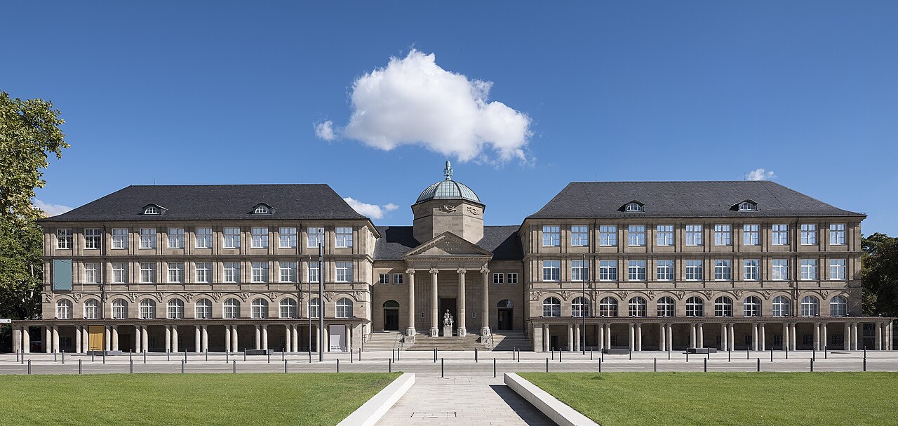 Ein sehr breites, klassizistisches Gebäude in hellem Gelb. In der Mitte tritt ein Portikus mit Säulen vor, über ihm sitzt eine kleine Kuppel mit Laterne. Zu beiden Seiten schließen lange, symmetrische Flügel mit dunklen Dächern an. Sie haben mehrere Reihen gleichmäßiger Fenster. Davor liegen Rasen und ein gerader Weg.