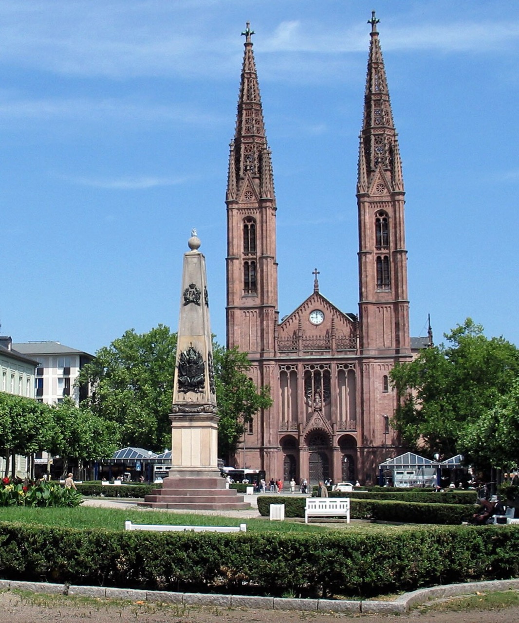 Eine neugotische Kirche aus rotem Stein mit zwei sehr hohen, schlanken Türmen. Zwischen den Türmen steht ein Giebel mit Uhr, darunter öffnet sich eine Vorhalle mit drei Bögen. Links davor steht ein Obelisk auf einem gestuften Sockel. Davor liegen eine Rasenfläche mit Hecke und Bäume.