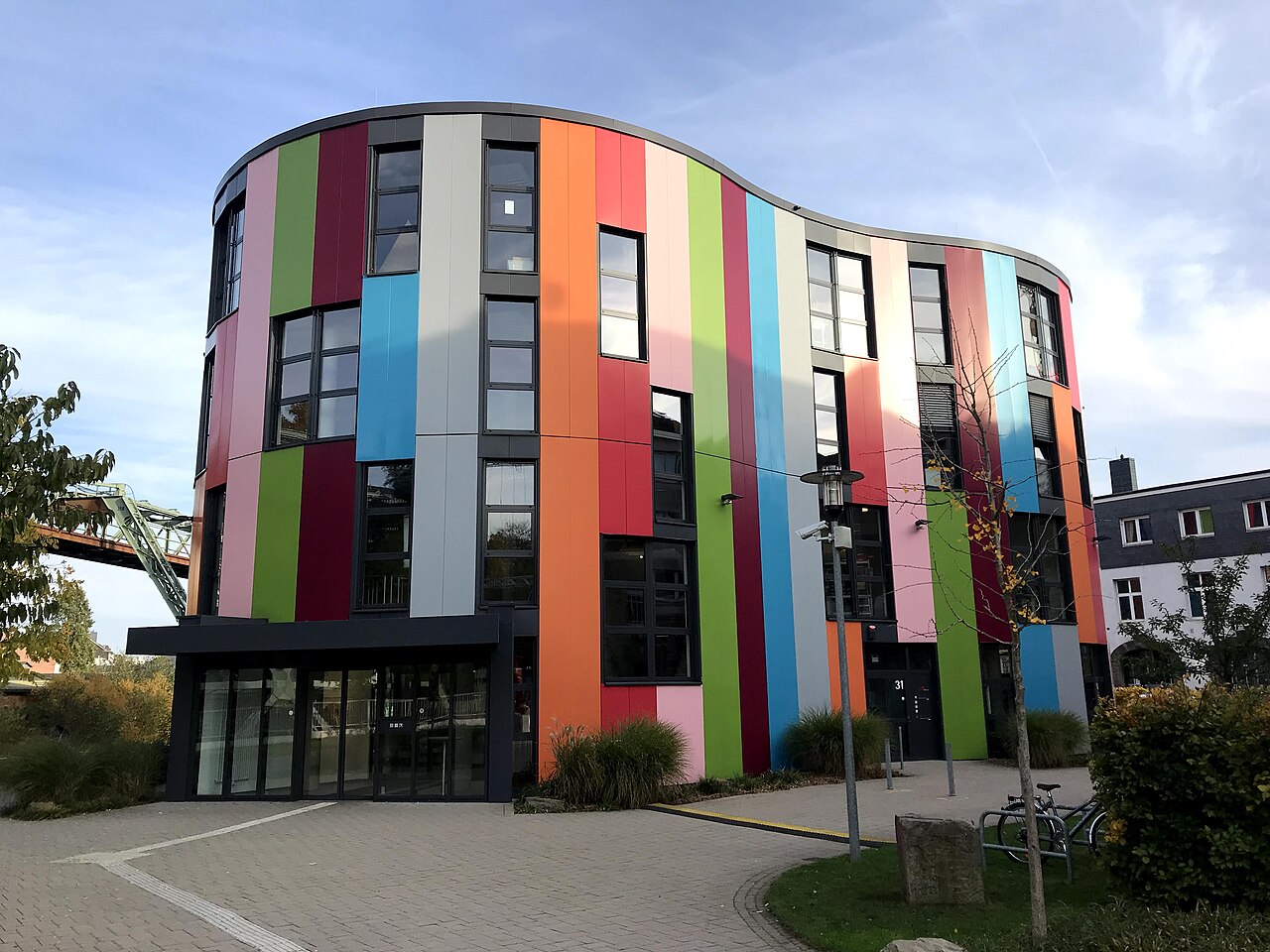 Ein runder, mehrgeschossiger Bau, dessen gebogene Fassade in senkrechte, farbige Streifen geteilt ist: Grün, Rosa, Grau, Orange, Blau und Rot. In jedem Streifen sitzt eine Reihe Fenster übereinander. Im Erdgeschoss liegt ein verglaster Eingang unter einem flachen Dach. Davor erstreckt sich ein gepflasterter Vorplatz mit Sträuchern.
